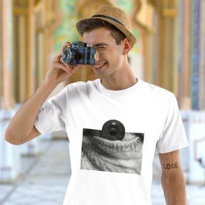 Man in a straw hat holding a vintage camera to his eye, wearing a white t-shirt with a black-and-white graphic of a human eye with a camera lens over the pupil. Photo taken in an ornate, archway-filled interior.