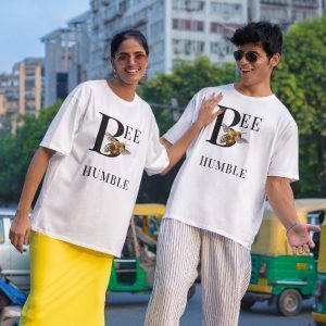 A happy couple stands playfully on a street curb. Both are wearing matching white oversized t-shirts with the text "BEE HUMBLE" and an illustration of a bee on the letter 'B'. The woman is wearing a yellow skirt and the man is wearing striped pants.