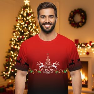 Smiling man wearing a red and black Merry Christmas T-shirt with reindeer and tree design, standing in a festive Christmas room.
